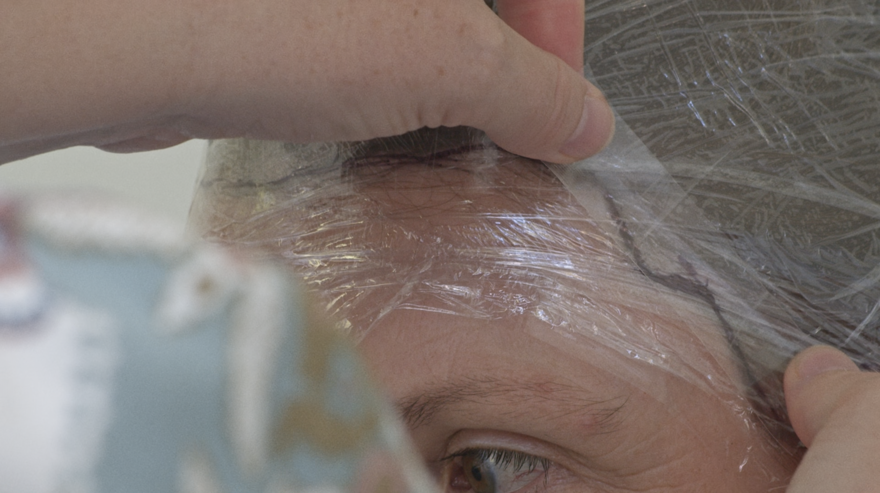 Film still shows a close up of a head with cling film covering the hair, a line drawn round the edge of the hairline and a pair of hands applying a strip of celeotape over the line.