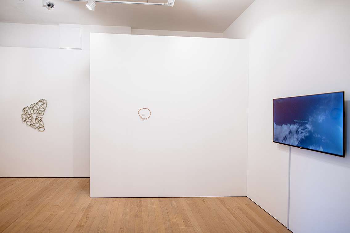 Image shows a gallery space with wooden floor, glass doors and white walls. On the left is a wall mounted ceramic, in the centre two ceramic hoops hang one on top of the other and to the right is a monitor showing a blue sky with light clouds and turquoise writing imposed over the top.