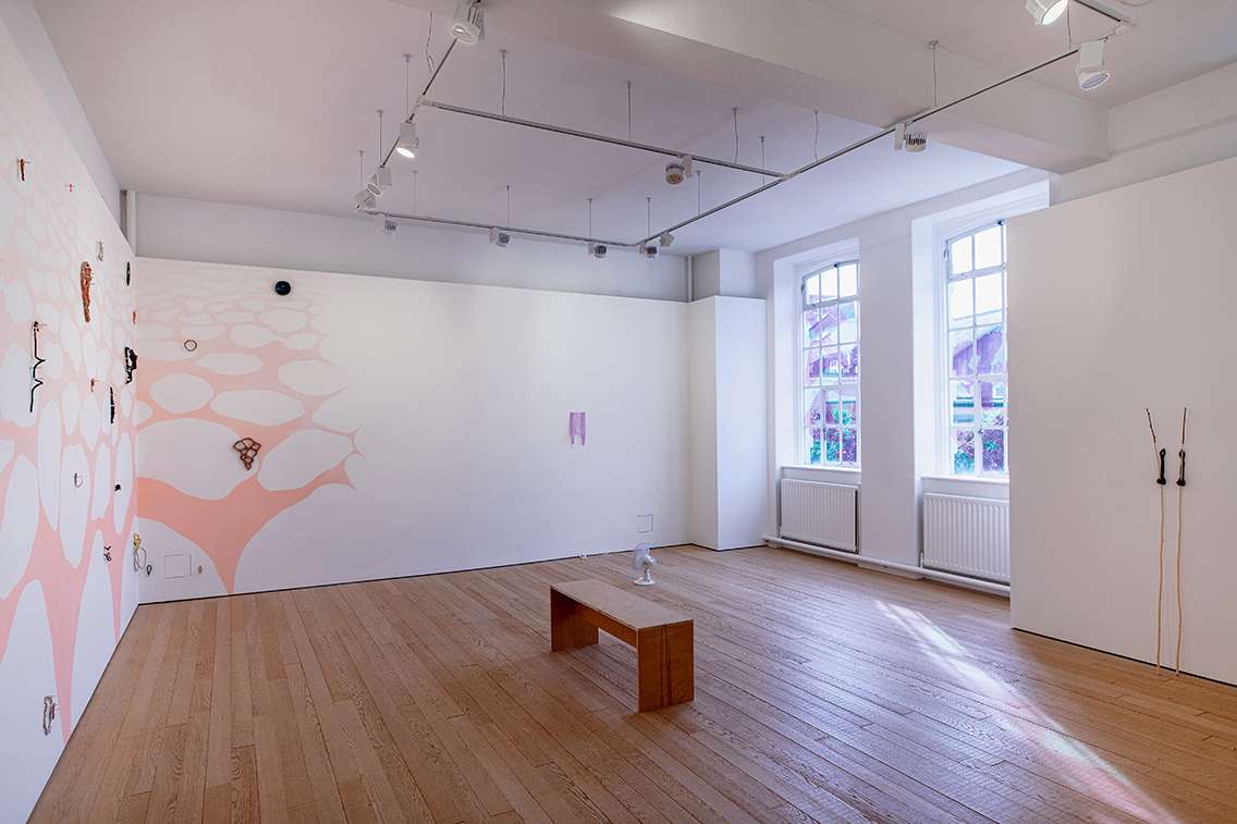 Image shows a gallery space with artworks installed, including a mural hung with ceramic drawings, a desk fan set on the floor rotating air that gently lifts a bag taped to the wall, windows with a repeating pink shape, and a pair of twigs held up right next to the wall. In the centre is a wooden bench.