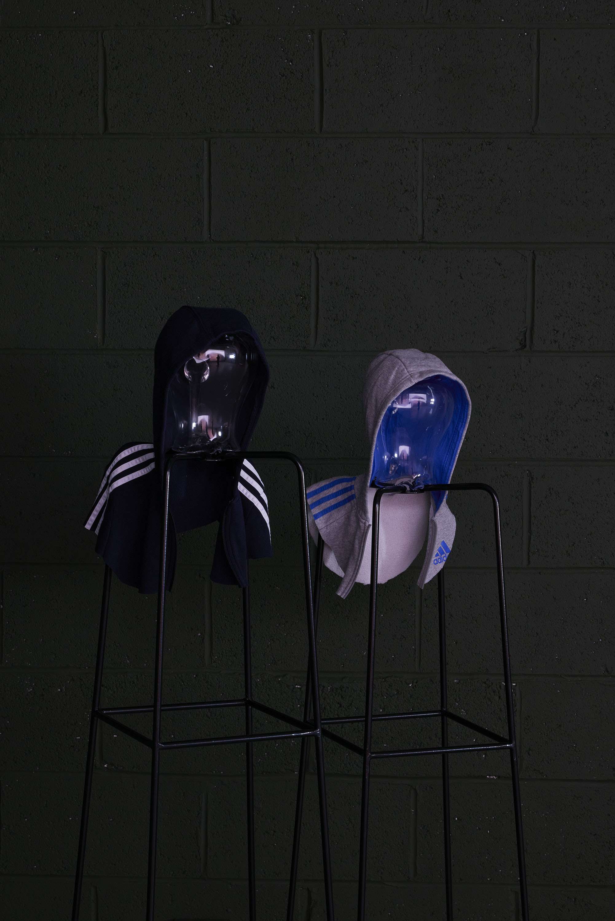 Image shows two figures suggested by glass heads, on black stands, wearing hoods, with a dark green wall behind. They are illuminated by light from a film playing in the space, which is reflected in the glass jug heads.