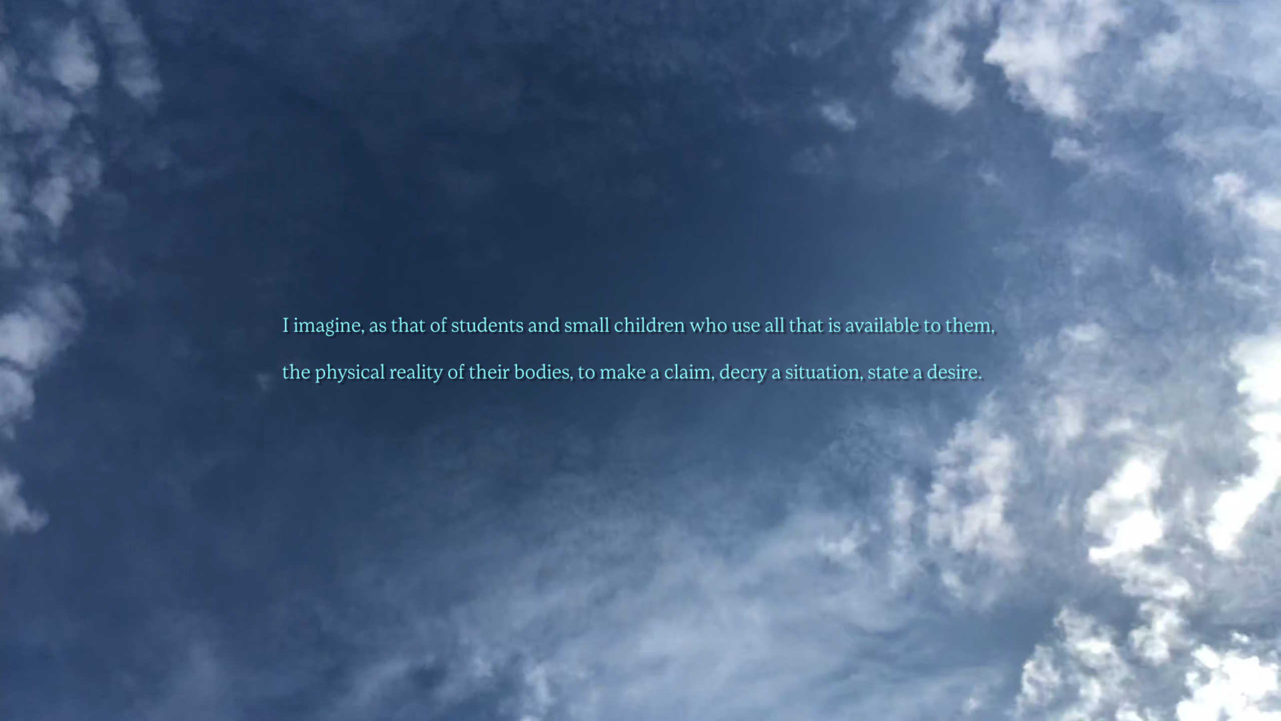 Image shows a screenshot of a blue sky with light clouds. Turquoise writing is imposed over the top, which reads, I imagine, as that of students and small children who use all that is available to them, the physical reality of their bodies, to make a claim, decry a situation, state a desire.