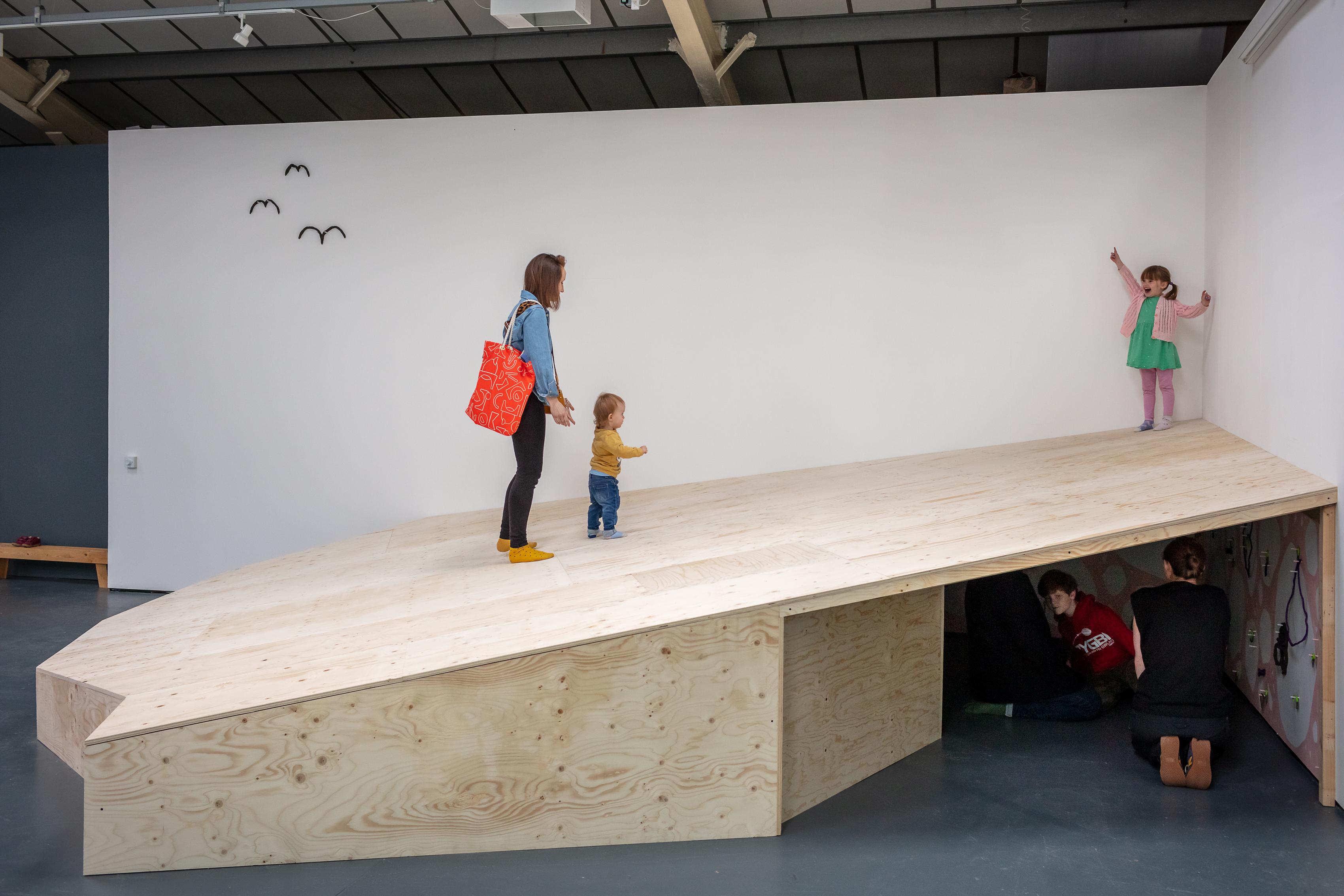 A large wooden ramp fills the corner of the gallery with three figures, an adult and two children on the ramp, and several figures visible in the opening underneath. The child at the top of the ramp points up, and further along the wall three birds appear to be suspended mid flight as if they were drawn there.