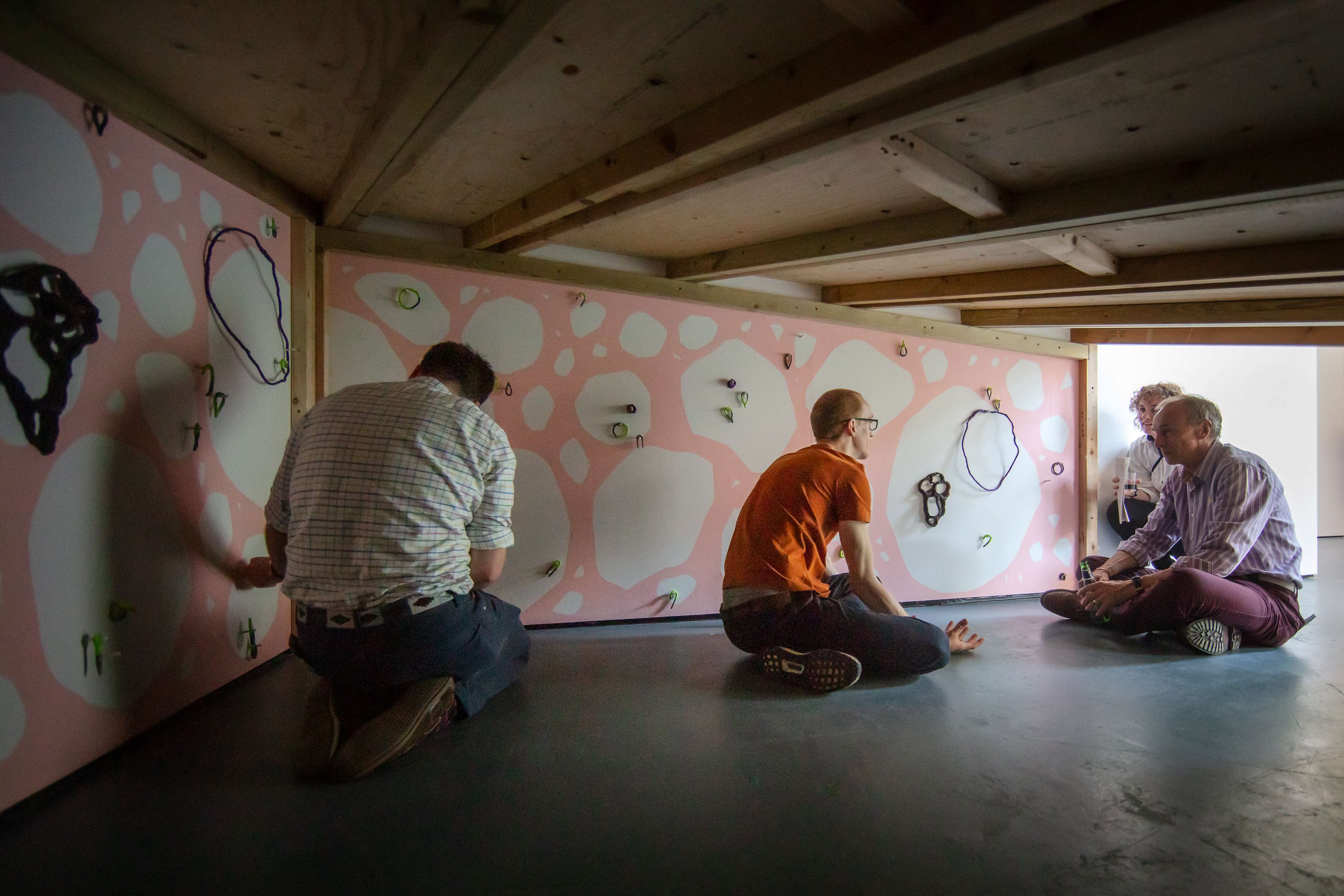 Four adults sit in a small space with a low wooden ceiling having a conversation. On the walls drawn shapes are hung from pegs protruding from a pink background with white boulders and rocks.