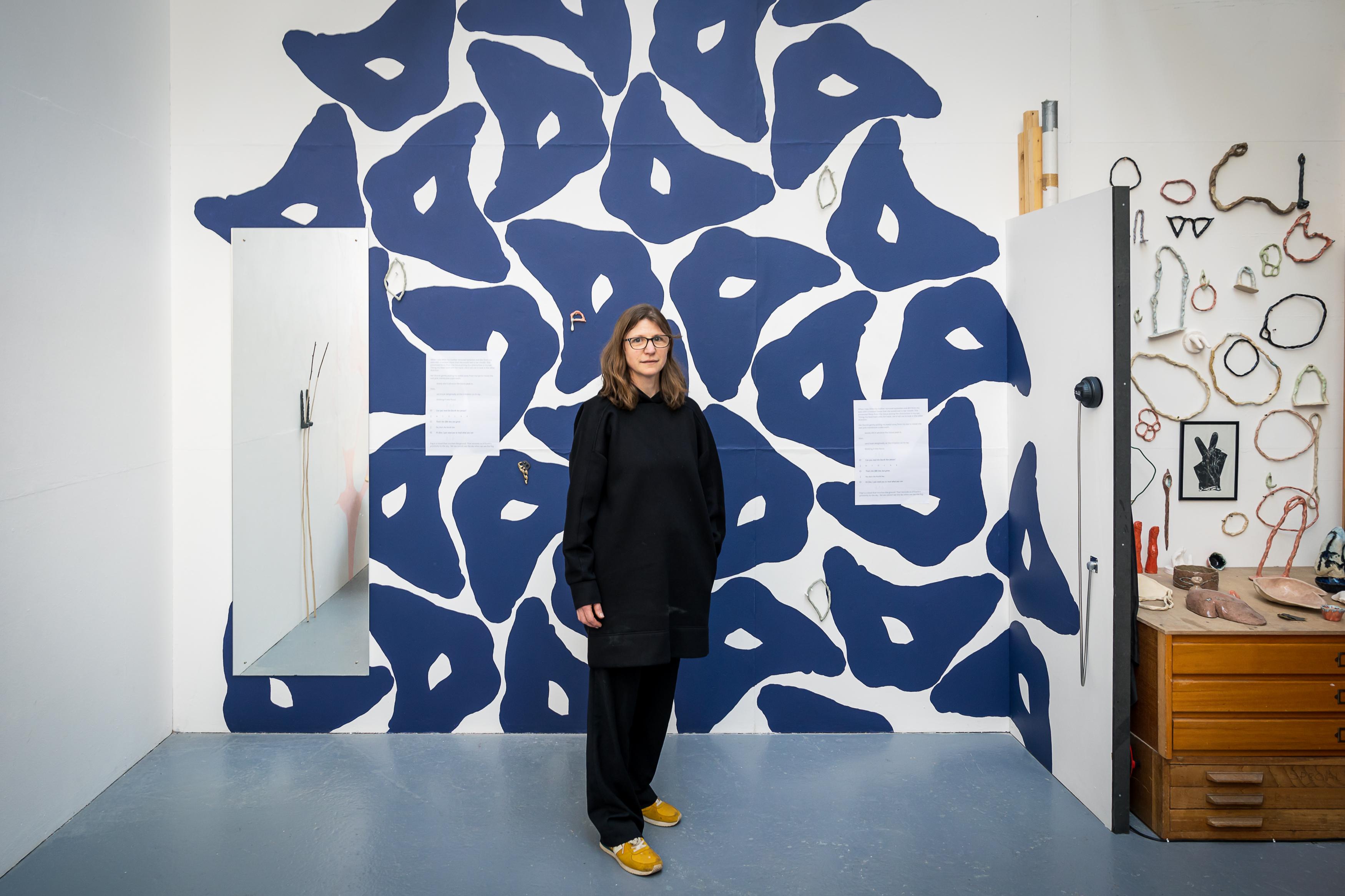 Image shows white woman dressed in black standing in front of a white wall with a dark blue shape repeating out of the top of the frame. There is a mirror in front of the mural showing a pair of twigs standing against the wall and ceramics on hte wall in the right of the frame.