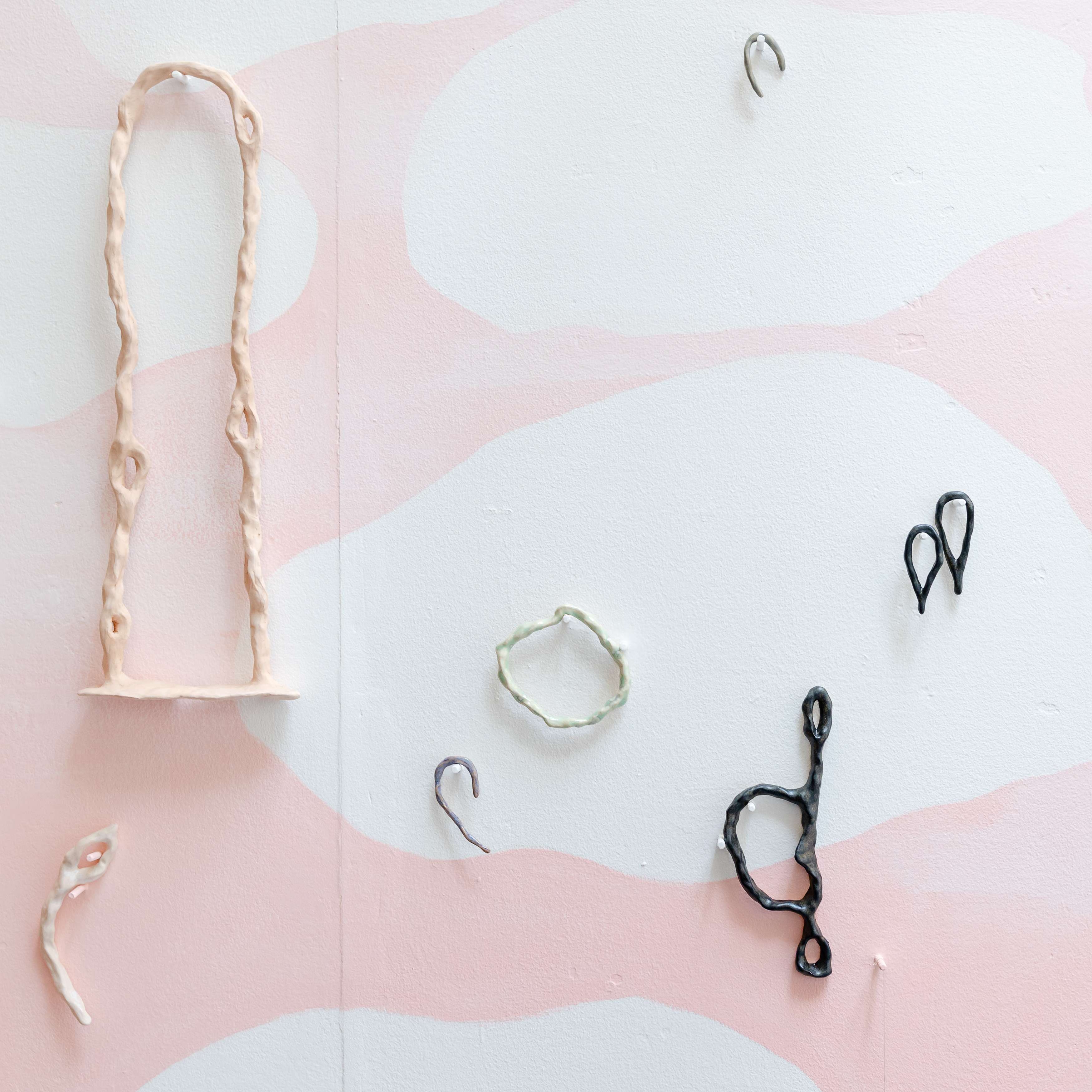 Image shows detail of a wall with white clouds retreating over a pink sky hung with drawn sculptural shapes. The objects include a stirrup, tears, hoops and ears.